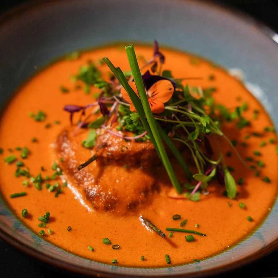 A rich tomato curry in a blue-grey bowl topped with herbs and an edible flower