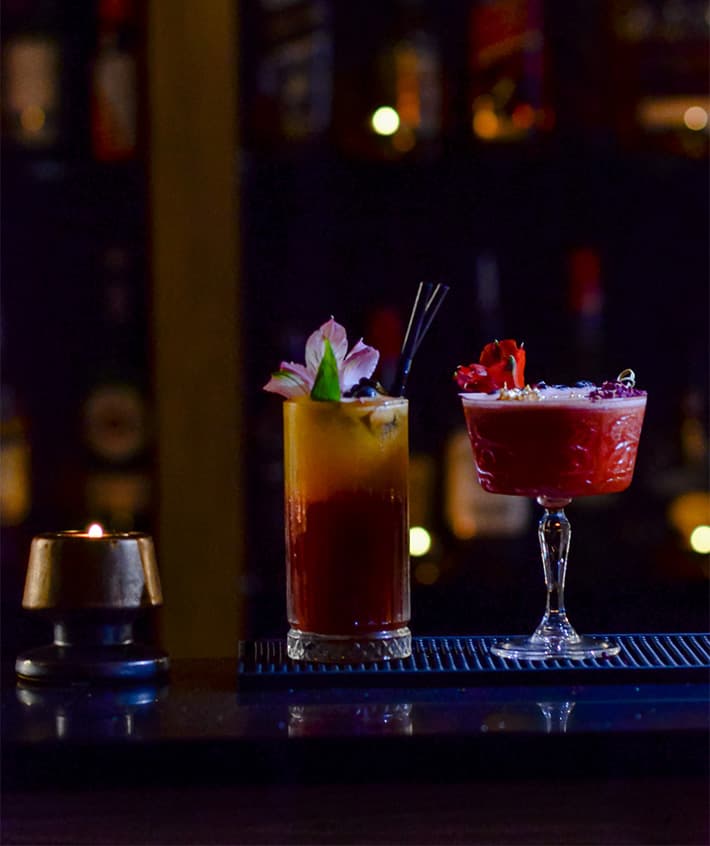Two cocktails on the bar, one amber and one deep red, lit from behind