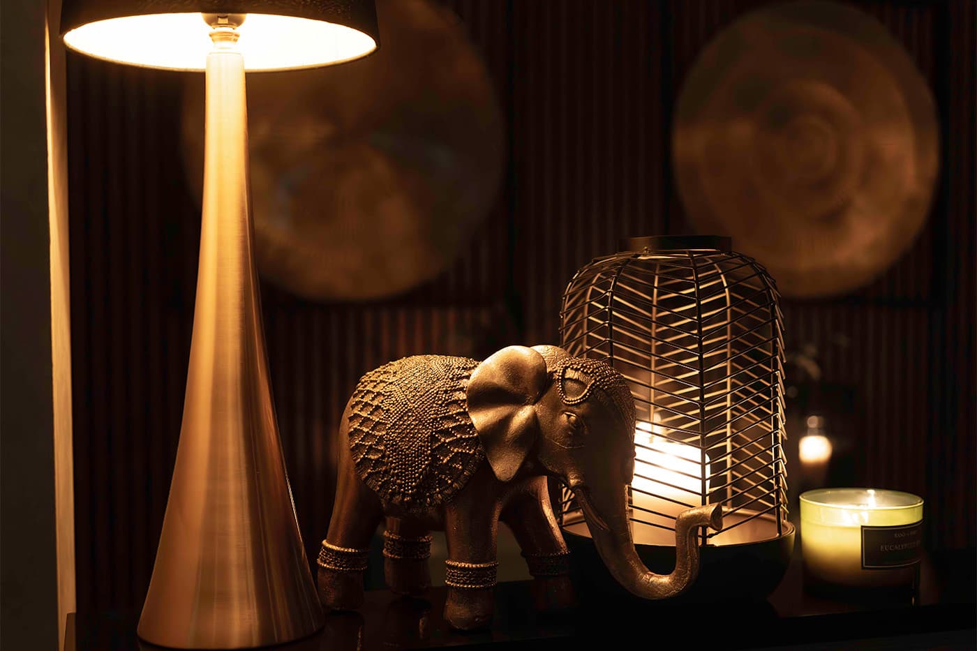 A brass elephant figure beside a lamp on a dark shelf