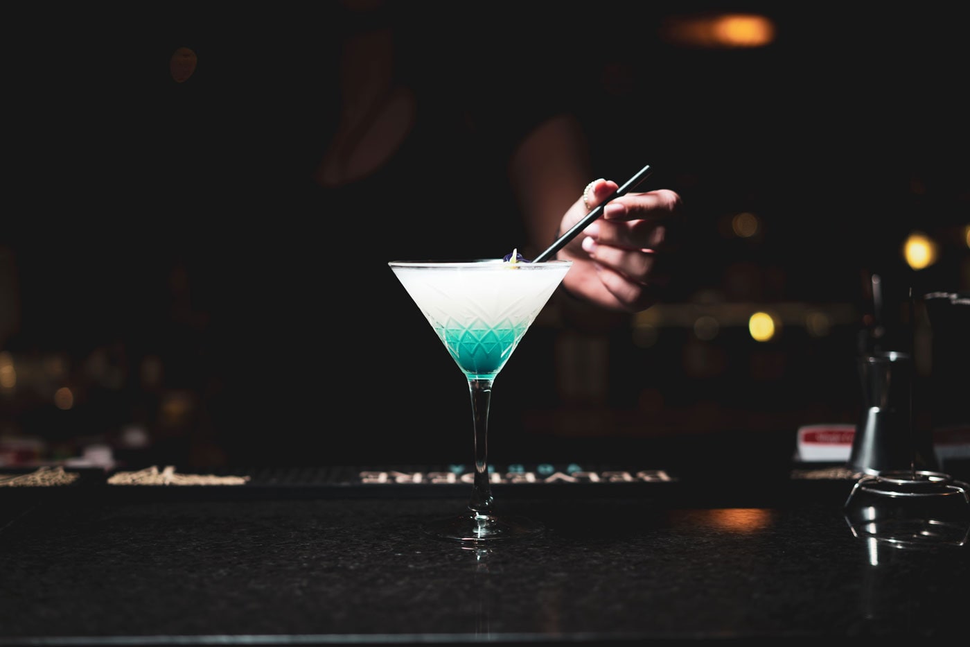 A pale blue martini on the bar with the room lights out of focus behind it