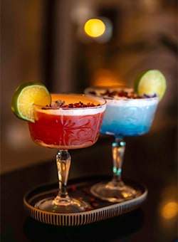 A pair of margaritas in patterned coupes, one red and one blue, with lime